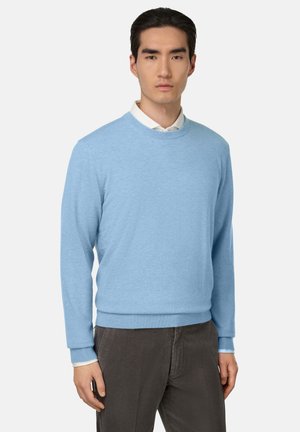 Un pull léger bleu clair à col rond fabriqué en tissu doux. Présente des poignets et un ourlet côtelés. Porté par-dessus une chemise blanche à col et associé à un pantalon sombre.