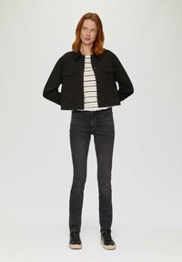 Zwarte cropped jack met knoopsluiting en twee voorzakken, gedragen over een gestreept shirt. Gecombineerd met zwarte slim jeans en zwarte sneakers.
