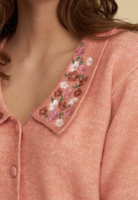 Miękki różowy sweter dzianinowy z kwiatowym haftem na kołnierzu, z brązowymi, białymi i lawendowymi kwiatami oraz zielonymi liśćmi.