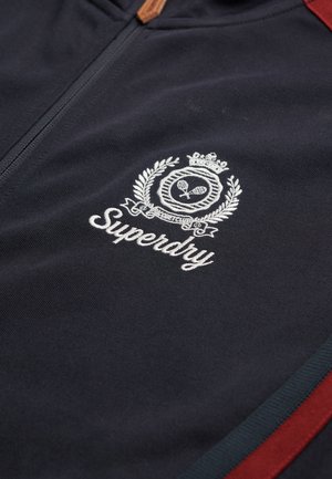 Marineblå lynlåsjakke lavet af tekstureret stof med et hvidt broderet logo med en krone og laurbærdesign, akkompagneret af røde striber.