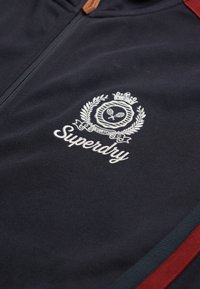 Marine zip-up jack met een gestructureerde stof, voorzien van een witte geborduurde logo met een kroon en laurierontwerp, geaccentueerd met rode strepen.
