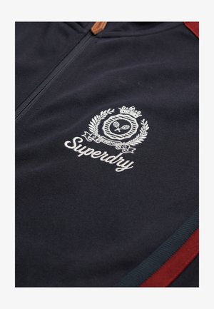 Marineblaues Zip-Jackett aus strukturiertem Stoff mit einem weißen gestickten Logo, das eine Krone und ein Lorbeerdesign zeigt, akzentuiert durch rote Streifen.