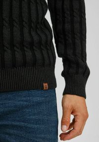 Black cable-knit sweater with ribbed hem and cuffs; features a small leather logo tag. Paired with textured blue denim jeans.