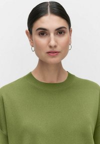 Grön stickad tröja med rund halsringning och ribbade detaljer, bärs av en modell med en subtil makeup-look.