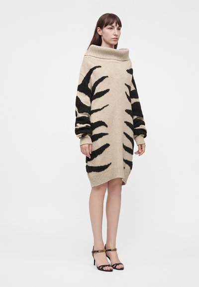 Γυναίκα στέκεται φορώντας μια μπεζ oversized πουλόβερ-φόρεμα με μαύρες αφηρημένες ρίγες και μαύρα πέδιλα με ψηλά τακούνια σε απλό λευκό φόντο.