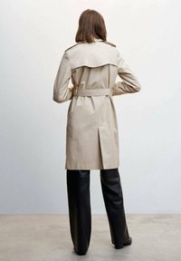 Beżowy płaszcz trench z paskiem w talii, epauletami na ramionach i wentylacją z tyłu, w zestawieniu z czarnymi skórzanymi spodniami. Gładki, lekki materiał.