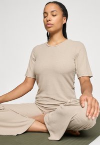 T-shirt beige à côtes et pantalon large assorti. Le tissu est doux avec une finition texturée. Le sujet est assis dans une pose méditative.