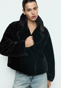 Sort faux fur frakke med bred krave, oversized pasform og knaplukning. Båret over lyseblå denim jeans. Glat tekstur.
