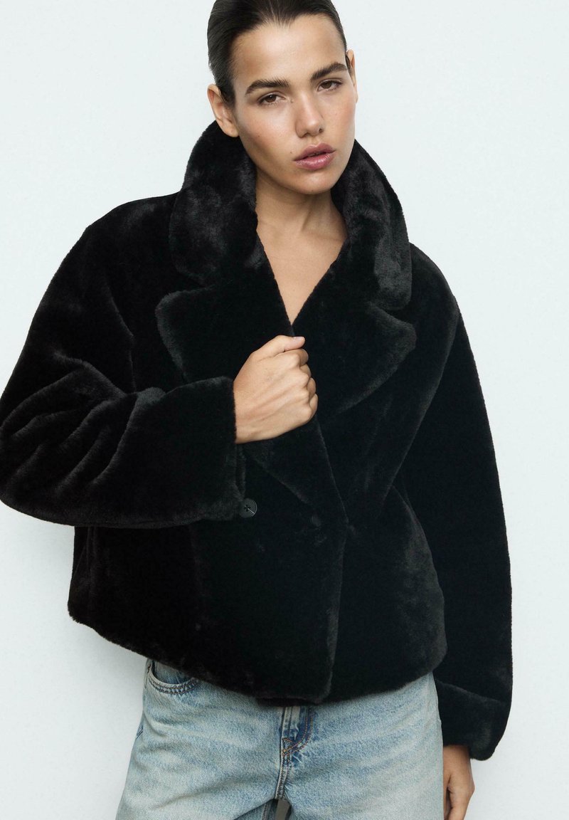 Sort faux fur frakke med bred krave, oversized pasform og knaplukning. Båret over lyseblå denim jeans. Glat tekstur.