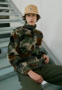 Fleecejakke i camouflage-mønster med nuancer af grøn, brun og creme, højt krave og lynlås, parret med grønne fleecebukser.