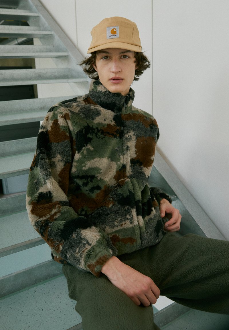 Fleecejakke i camouflage-mønster med nuancer af grøn, brun og creme, højt krave og lynlås, parret med grønne fleecebukser.