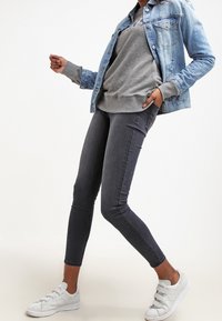 Chaqueta de mezclilla en azul claro, sudadera gris debajo, jeans skinny gris oscuro y zapatillas blancas sin cordones. Texturas suaves y diseño ajustado.