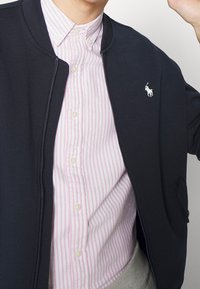 Námořnická zipová bunda přes růžovou a bílou pruhovanou košili s knoflíky, která má na hrudi malé vyšívané logo a texturovanou tkaninu.