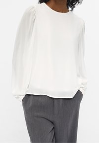 Blouse blanche et transparente à manches longues, avec des détails froncés aux épaules et un col arrondi ; associée à un pantalon gris ajusté.