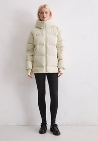 Gewatteerde jas in lichtbeige, met een hoge kraag, volledige rits en ribgebreide manchetten. Gecombineerd met zwarte leggings en stevige zwarte schoenen.
