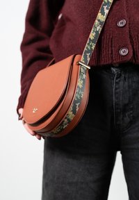 Braune Leder-Crossbody-Tasche mit runder Form, ausgestattet mit einem gemusterten Schulterriemen in dunklen Farben, verziert mit floralen und tierischen Motiven.