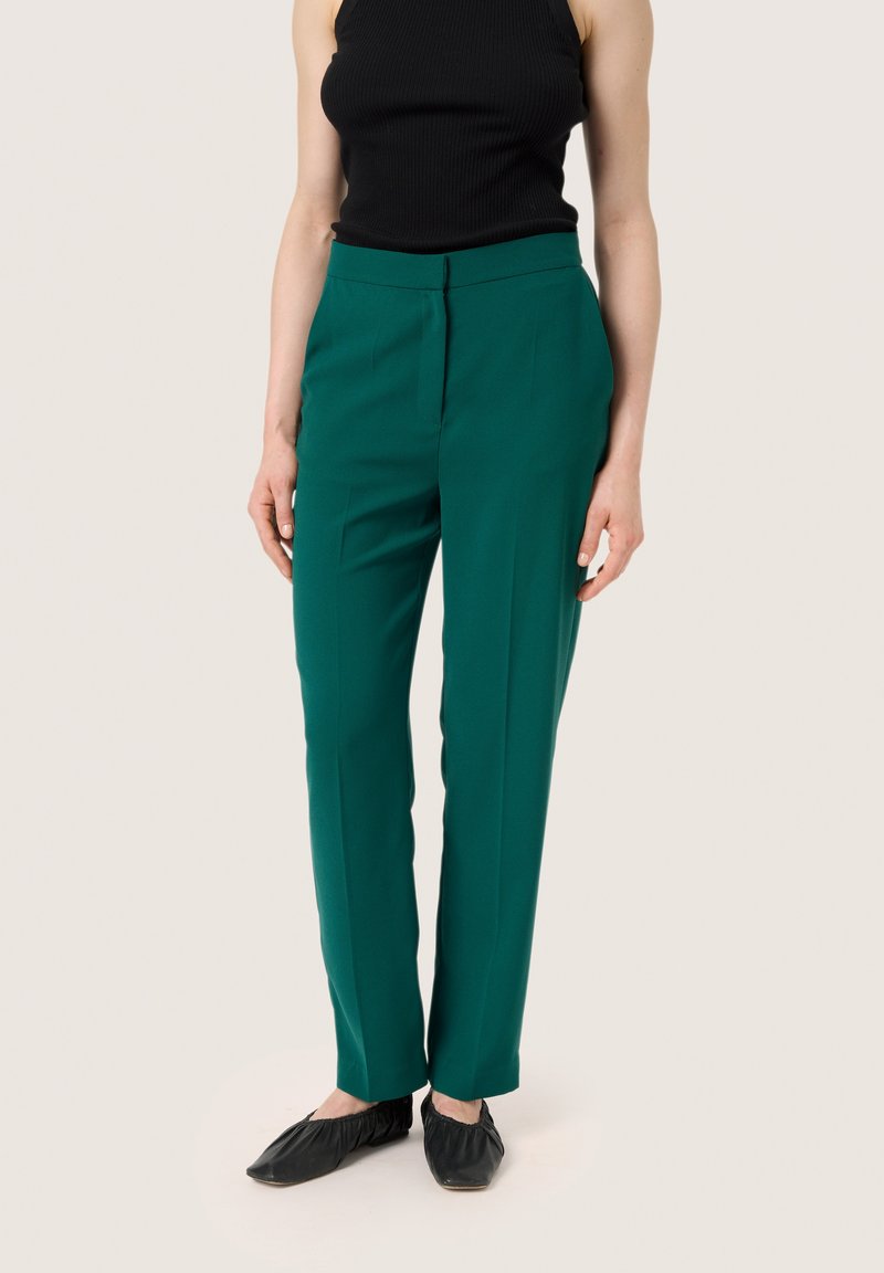 Zelené kalhoty na míru se hladkým povrchem, s páskem střední výšky a slim fit. Kombinováno s černými nazouvacími botami.