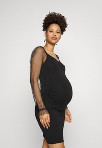 Vestido de maternidade preto com silhueta ajustada, apresentando mangas em rede transparente e um corpete franzido, projetado para conforto e estilo.