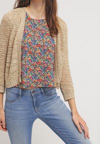Beige stickad cardigan över en blus med blommönster i blå, röd och grön nyanser. Ljust blå jeans fulländar outfiten.