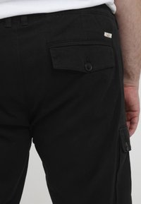 Schwarze Cargo-Hosen mit einer seitlichen Tasche und einem geknöpften Deckel. Glatte Textur und dezente Steppnähte. Das Etikett trägt die Aufschrift "BUENA."