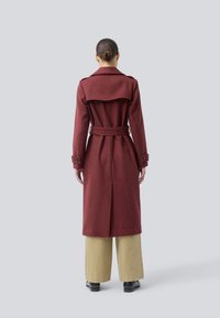 Kvinna står med ryggen mot kameran iförd en lång burgunderfärgad kappa med bälte, beige vida byxor, svarta skor och med håret i en knut.