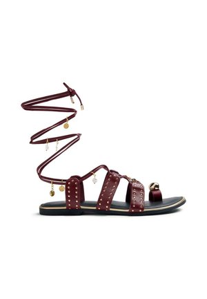 Sandalia plana burdeos con tres tiras tachonadas, lazo para el dedo y largas cintas para atar al tobillo adornadas con dijes de oro y perlas.