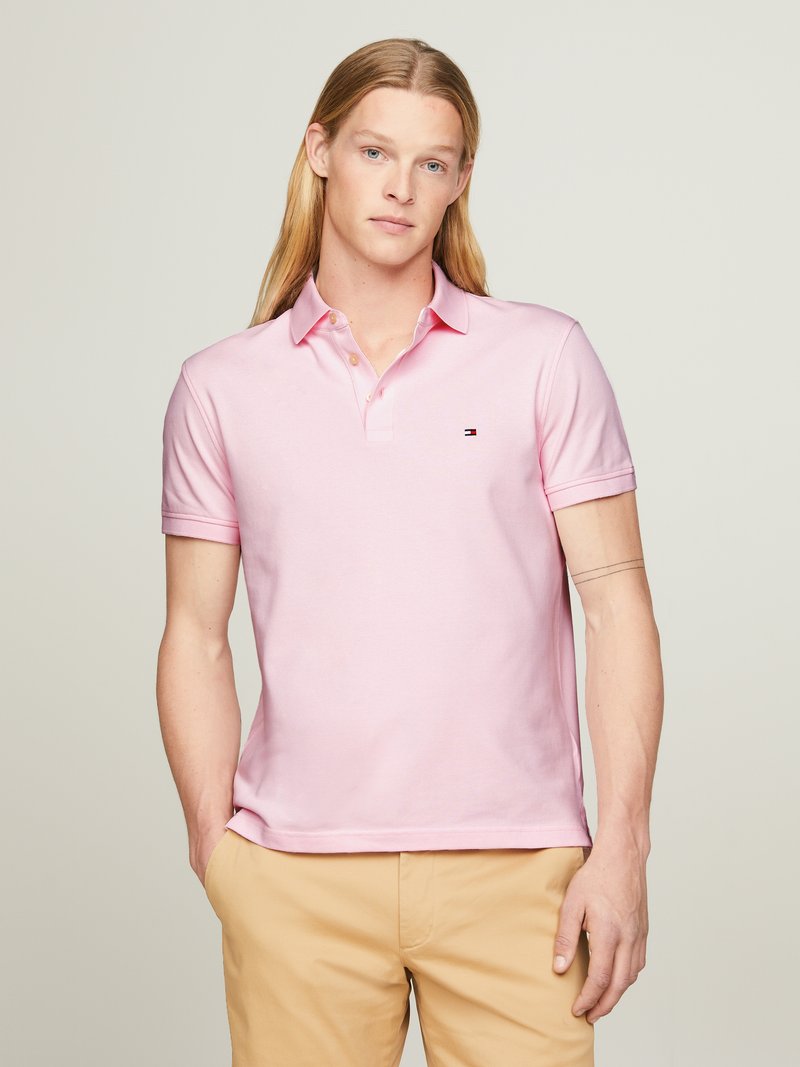Polo shirt rosa claro com colarinho e mangas curtas, apresentando um pequeno logótipo no lado esquerdo do peito. Feita de um tecido suave e texturizado.