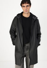 Black waterproof coat with a hood, featuring a front zip, side pockets, and a smooth texture. Worn over a black sweater and gray jeans.