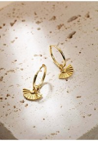 Pendientes de aro de oro que presentan un colgante en forma de abanico con una superficie texturizada y líneas radiales. Mostrados sobre un fondo de piedra de color claro.