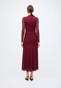 Burgundy long-sleeve dress with ruched detailing at the waist. Sheer sleeves, flowing skirt, and a high neckline. Back zipper closure.