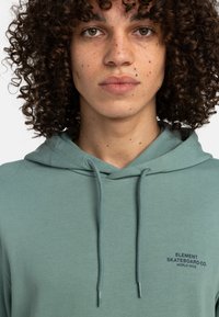 Sudadera verde menta con cordón, que presenta un pequeño logo negro en el lado izquierdo del pecho. Textura suave y corte estándar.