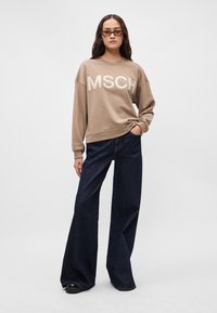 Καφέ φούτερ με το "MSCH" σε κρεμ γράμματα, συνδυασμένο με φαρδιά σκούρα μπλε τζιν. Το μοντέλο φοράει στυλάτα γυαλιά ηλίου και στέκεται με τα χέρια στις τσέπες.