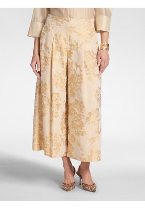 Pantaloni beige a gamba larga con motivo floreale dorato, abbinati a décolleté con punta decorata e un bracciale a polsino dorato al polso destro.