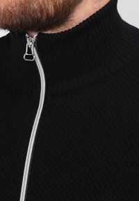 Schwarzer Reißverschluss-Pullover mit einem strukturierten Muster, hohem gerippten Kragen und silbernem Reißverschlussdetail. Weicher Stoff, sichtbare Nähte und Kragenstruktur.