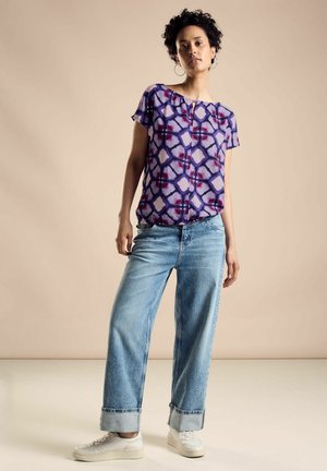 Lila gemusterte Bluse mit kurzen Ärmeln, kombiniert mit hellblauen weiten Jeans und weißen Turnschuhen. Der Stoff wirkt leicht und strukturiert.
