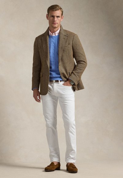 Muž v hnědém blejzru, modrém svetr s copánkovým vzorem, bílých kalhotách a hnědých mokasínách, stojící před jednobarevným béžovým pozadím.