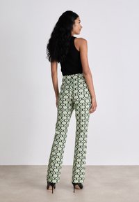 Gemusterte Hosen mit hoher Taille präsentieren einen grünen Grundton und geometrische Formen in Schwarz und Weiß, kombiniert mit einem schwarzen taillierten Oberteil und Absatzschuhen.