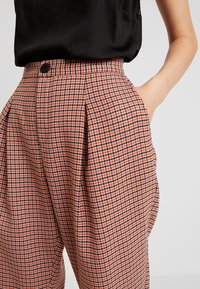 Rutig byxa med en övervägande röd och svart design, hög midja, sidfickor och knappstängning. Slät textur.