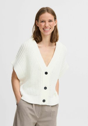 Cárdigan blanco de punto con un escote en V profundo, mangas cortas y botones negros. Llevado sobre pantalones grises a rayas. Tejido texturizado.
