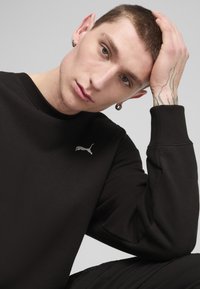 Sudadera negra con puños y cuello acanalados, que presenta un pequeño logo gris de Puma en el pecho, llevada por una persona con cabello corto y pendientes.