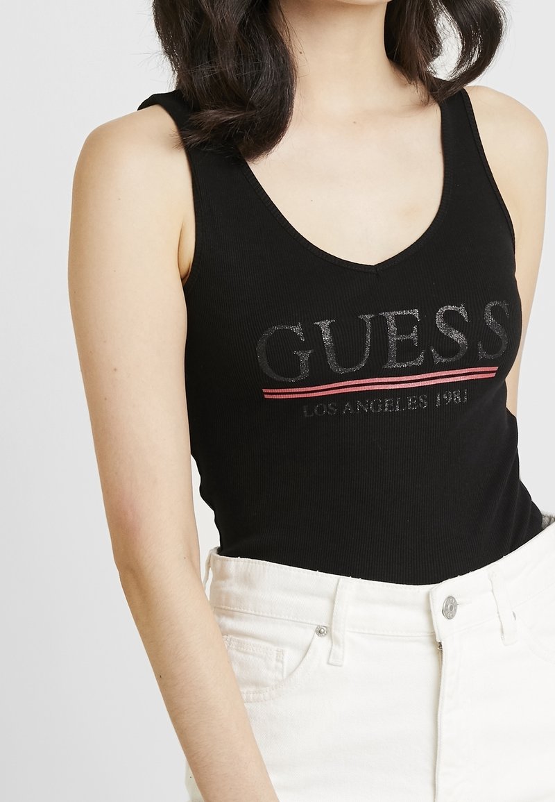 Top senza maniche nero a coste con scollo a V, caratterizzato da un audace logo "GUESS" in grigio metallico e rosso. Indossato con jeans bianchi a vita alta.