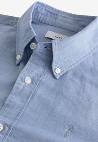 Hellblaues Baumwollhemd mit einem Knopfkragen, weißen Knöpfen und einem auf der Brust gestickten Logo. Strukturierter Stoff mit dezenten Mustern.