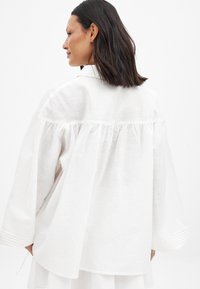Baltas lina blūze ar brīvu piegriezumu, savāktas aizmugures dizainu un garām piedurknēm ar smalkām tekstūras akcentiem.