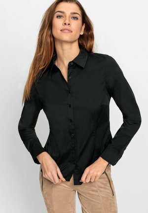 Jonge vrouw met lang bruin haar, gekleed in een zwart overhemd en beige broek, poseert tegen een effen lichte achtergrond.
