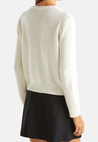 Weißer gestrickter Pullover mit geripptem Saum und Bündchen, runder Ausschnitt und strukturiertem Finish. Getragen über einem schwarzen Rock.