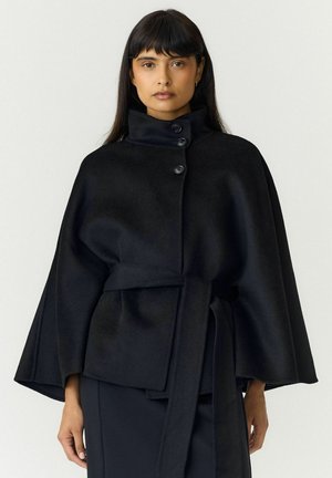 Mujer vestida con un abrigo capa negro abotonado, con cuello alto y cinturón atado, de pie frente a un fondo claro y liso.