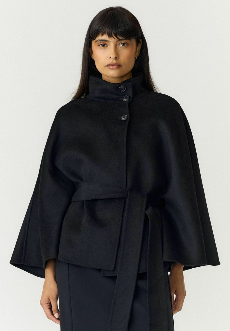 Femme portant un manteau cape noir boutonné avec un col montant et une ceinture nouée, debout devant un fond clair uni.