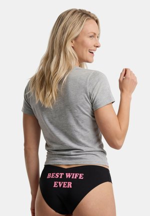 Blondynka uśmiecha się przez ramię, ubrana w szarą koszulkę i czarne majtki z różowym napisem "NAJLEPSZA ŻONA NA ŚWIECIE" na plecach.