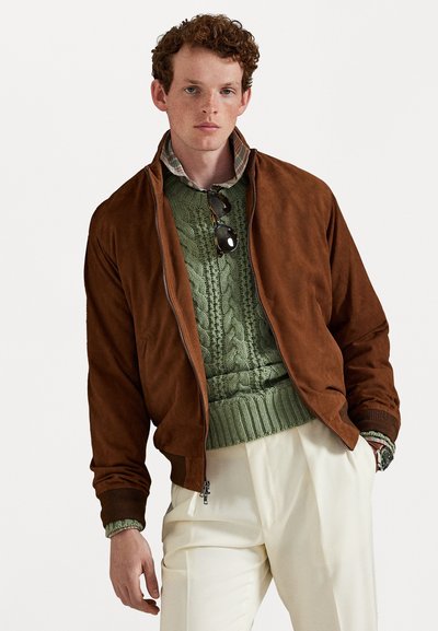 Braune Wildleder-Bomberjacke mit einem Reißverschluss, getragen über einem grünen Kabelstrickpullover. Das Modell trägt cremefarbene Hosen und Sonnenbrillen.