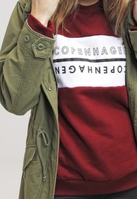 Oliwkowa parka na burgundowy sweter z białą taśmą z napisem "COPENHAGEN" wydrukowanym na czarno, o luźnym kroju i teksturowanej tkaninie.